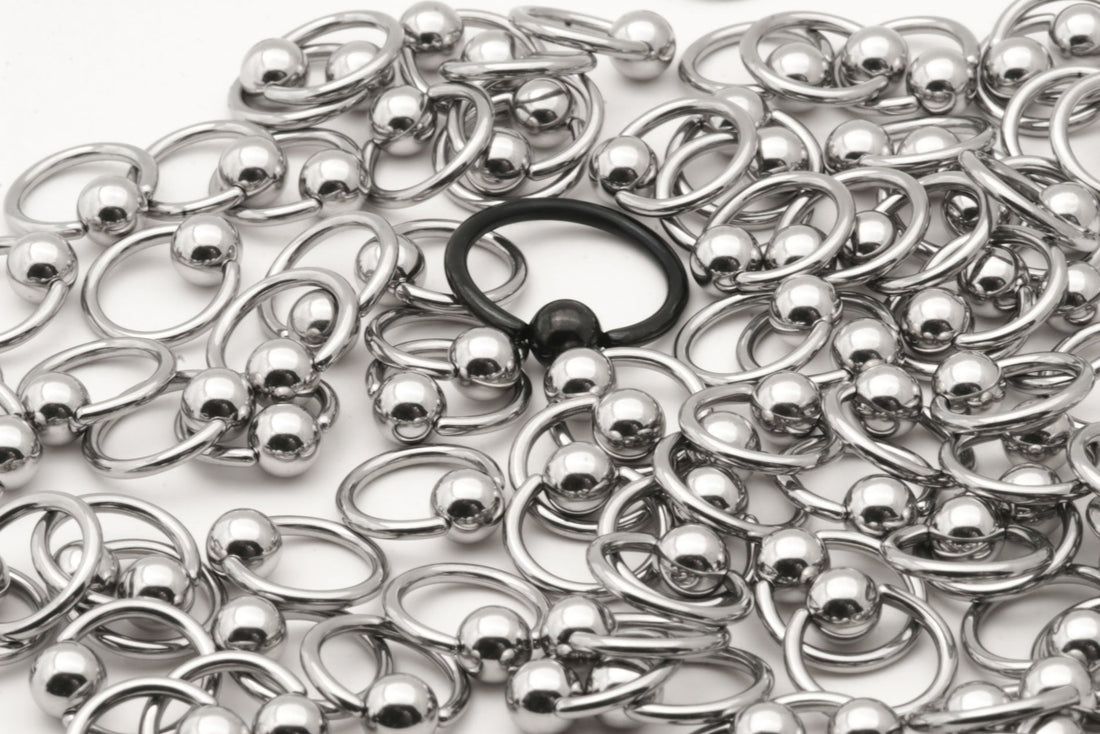 A pile of surgical steel captive bead rings and one singular black captive bead ring pictured against a white background.