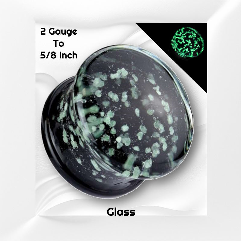 One of a pair of glass glow in the dark gauges with green glowing speckles embedded in the glass plug pictured on a white wavy background with black text stating the sizes available of this gauge earring.